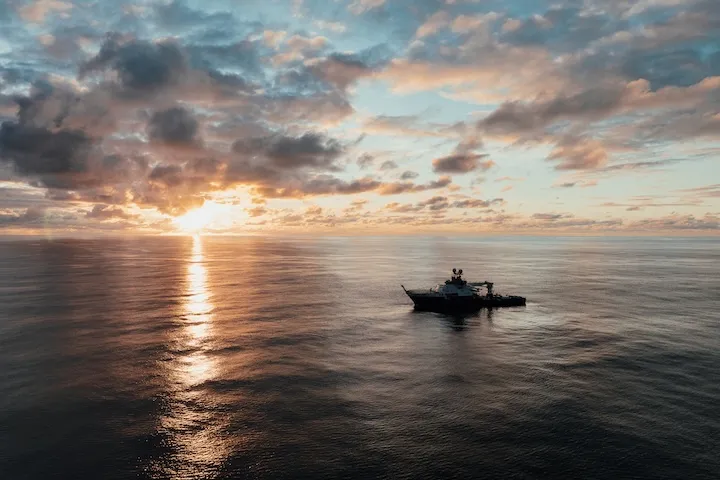 Research vessel sailing on calm ocean waters during a colorful sunset with a cloudy sky.