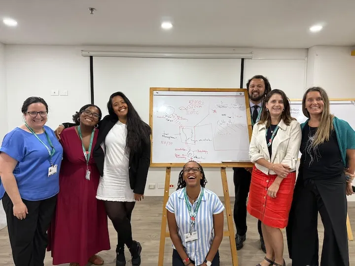 Seven smiling people posing in front of a whiteboard with diagrams and notes in a classroom setting.