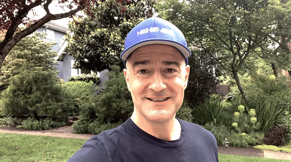 Man wearing a blue 1-800-GOT-JUNK? cap and navy shirt smiling outdoors with green trees and plants in the background.