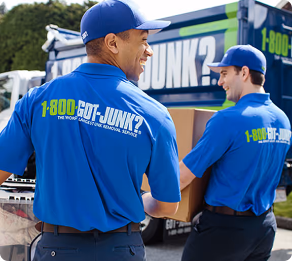 Two workers in blue 1-800-GOT-JUNK? uniforms carrying a cardboard box near a 1-800-GOT-JUNK? truck.