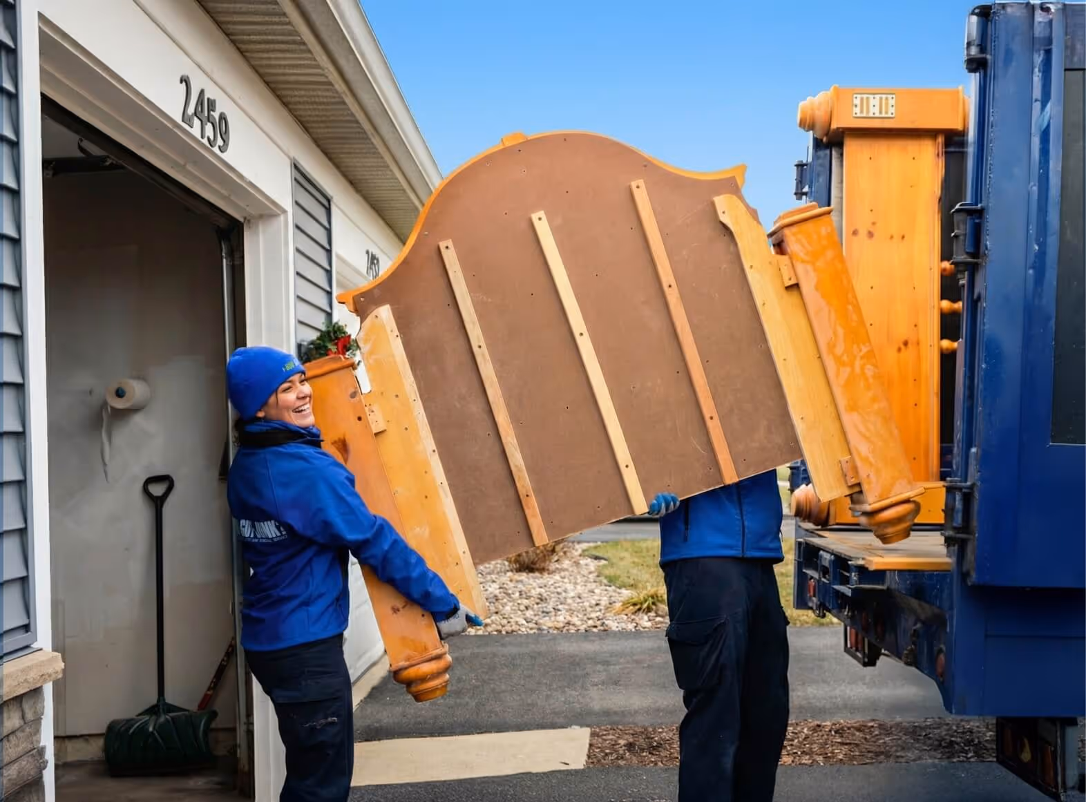 Two movers in blue 1-800-GOT-JUNK? jackets carrying a wooden bed frame from a house to a truck.