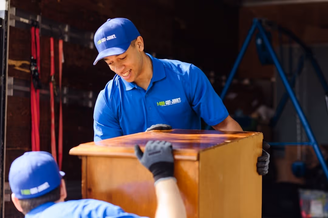 Two men in blue uniforms with 1-800-GOT-JUNK? logos lifting a wooden dresser in a garage or storage area.