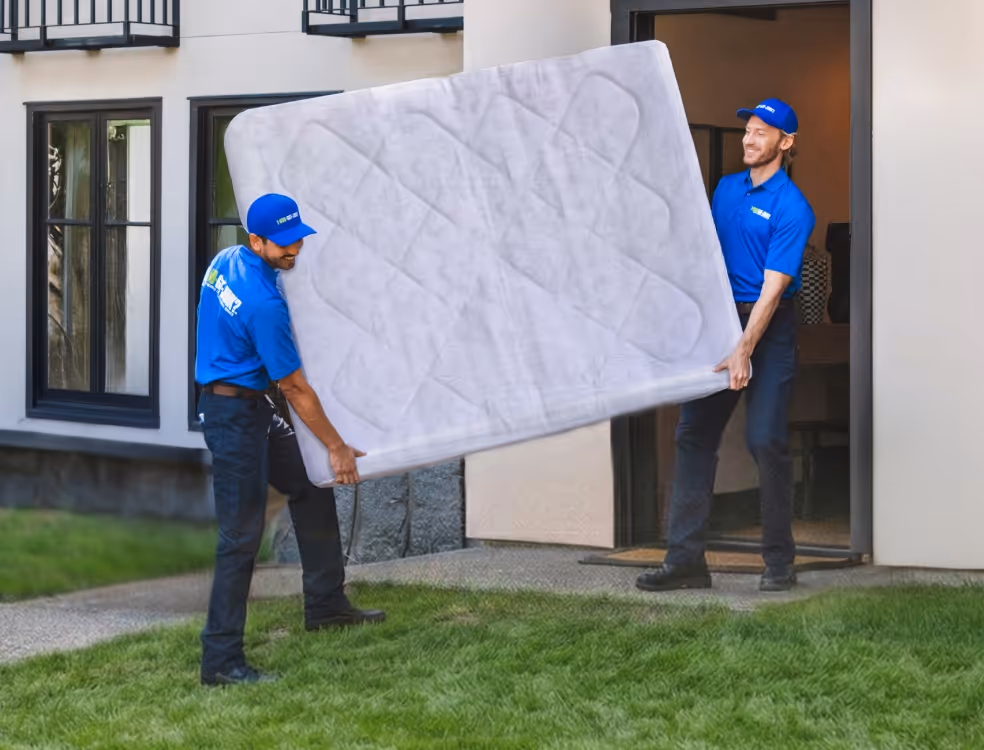 Two movers in blue uniforms carrying a mattress out of a house.