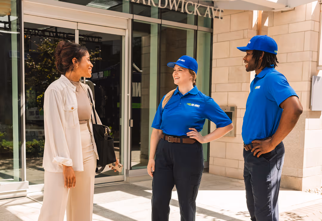 Two service workers in 1-800-GOT-JUNK? blue uniforms smiling and talking with a person in beige clothing outside a building entrance.