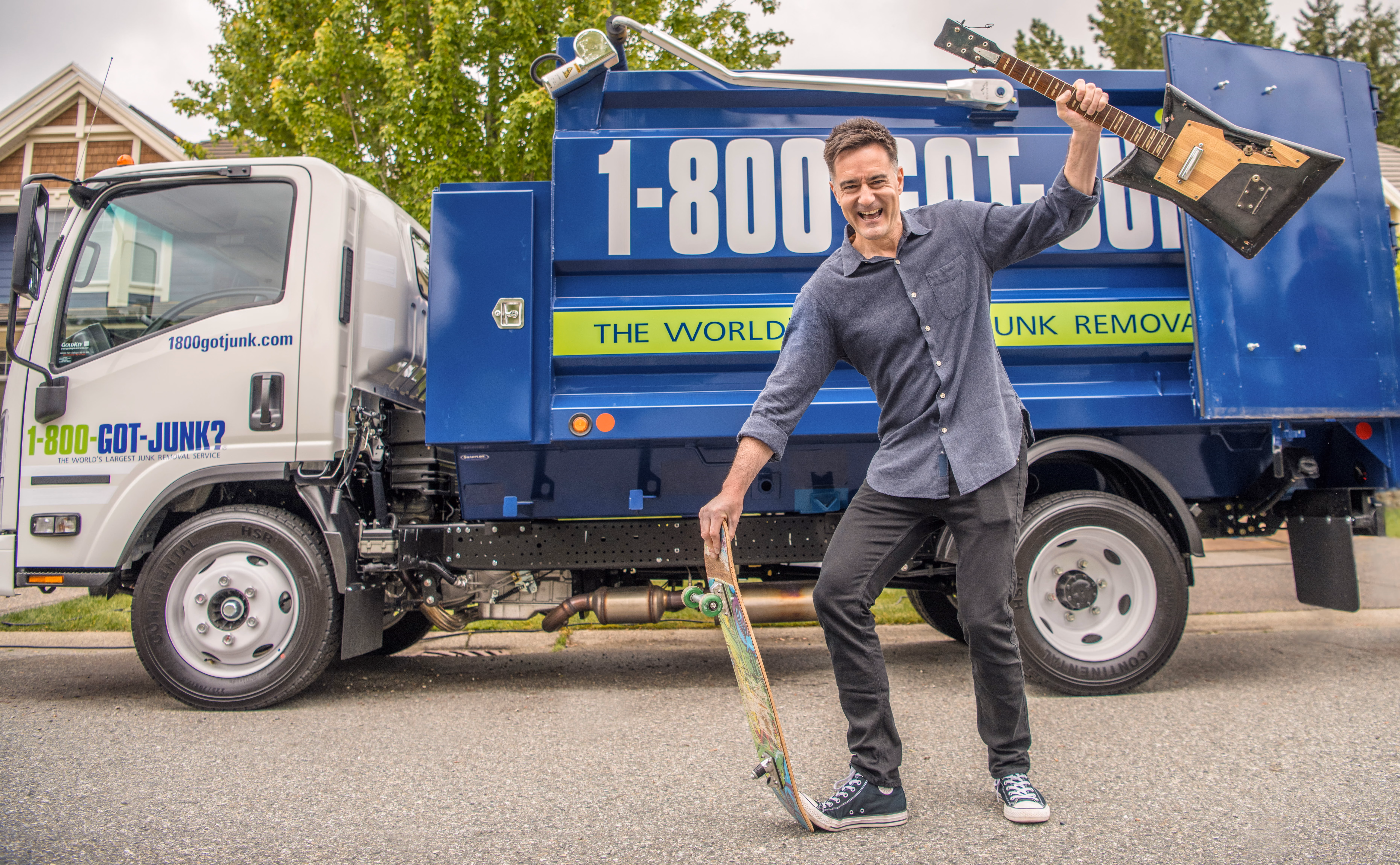 Man smiling and holding a skateboard and a guitar in front of a 1-800-GOT-JUNK? truck.