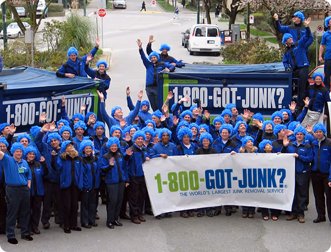 Group of 1-800-GOT-JUNK? employees wearing blue uniforms and blue wigs, standing around two trucks and holding a banner with the company's name and slogan.