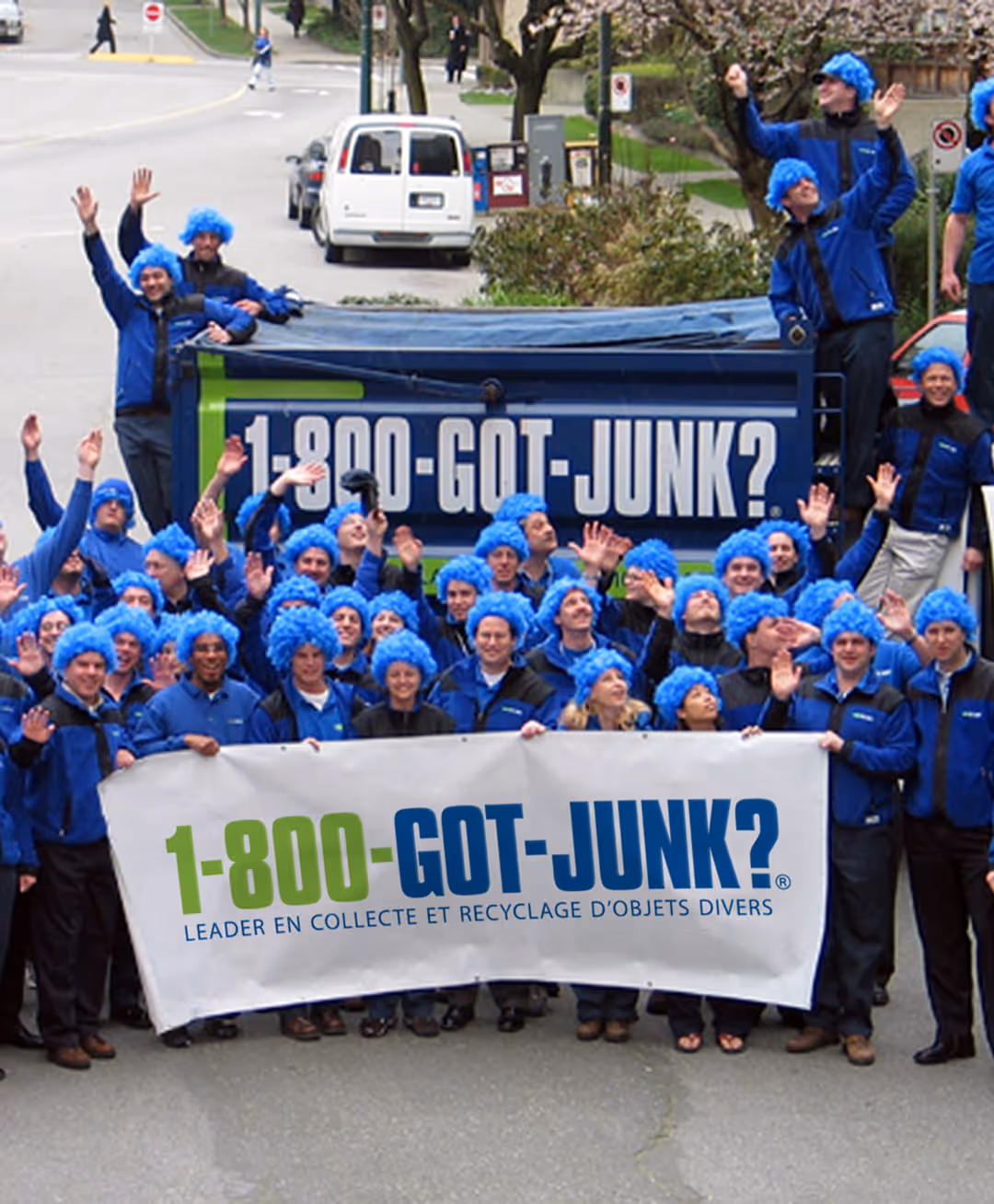 Group of people wearing blue wigs and jackets holding a banner with 1-800-GOT-JUNK? logo in front of a truck with the same logo.