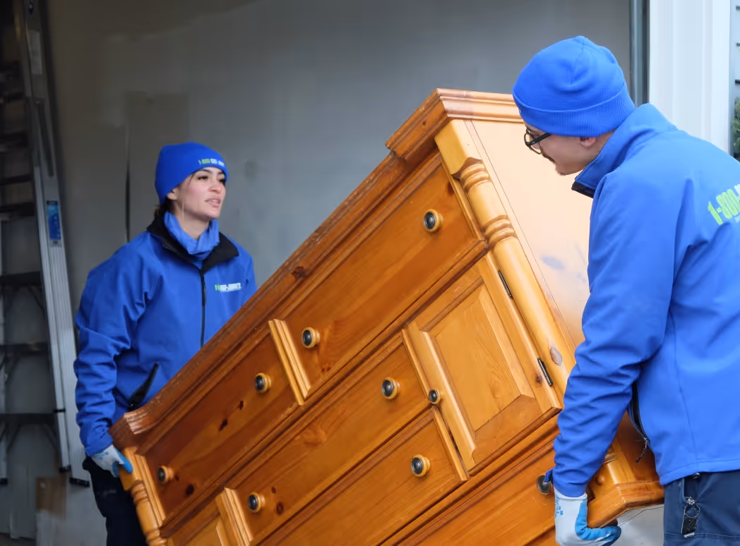 Two movers in blue 1-800-GOT-JUNK? jackets and beanies carrying a large wooden dresser.