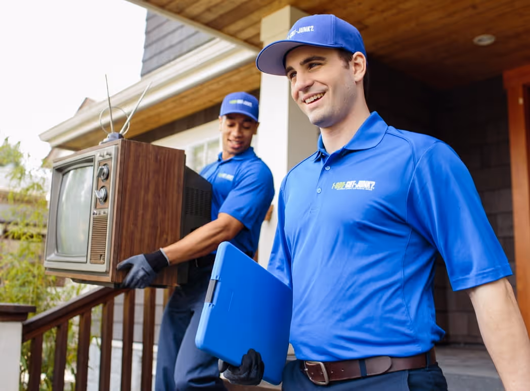 Two workers in blue 1-800-GOT-JUNK? uniforms carrying an old television and a blue clipboard outside a house.