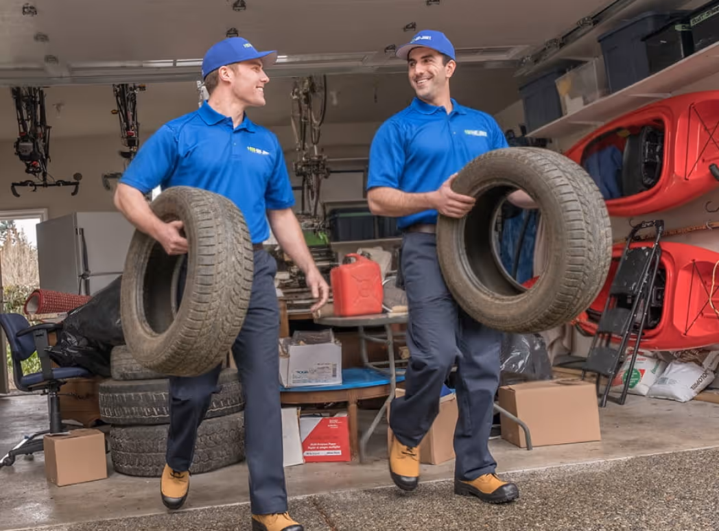 Two workers in blue 1-800-GOT-JUNK? shirts and caps carrying tires in a garage filled with boxes, kayaks, and bicycles.