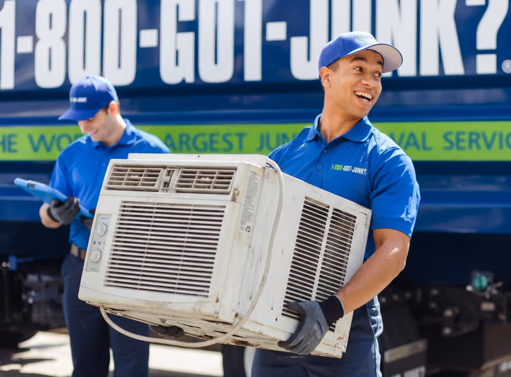 Two junk removal workers in blue uniforms handling an old air conditioning unit in front of a truck with 1-800-GOT-JUNK? branding.