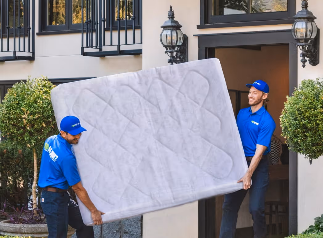 Two men in blue 1-800-GOT-JUNK? uniforms carrying a large mattress outside a building entrance.