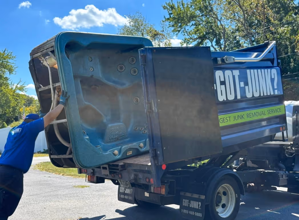 Worker loading a large hot tub into a 1-800-GOT-JUNK? junk removal truck on a sunny day.