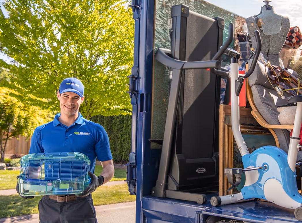 Smiling man in a blue 1-800-GOT-JUNK? uniform holding a small pet cage near a blue truck loaded with household items.