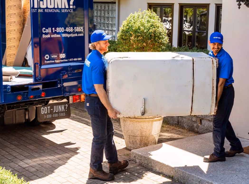 Two men in blue uniforms carrying an old refrigerator from a house to a 1-800-GOT-JUNK? truck.