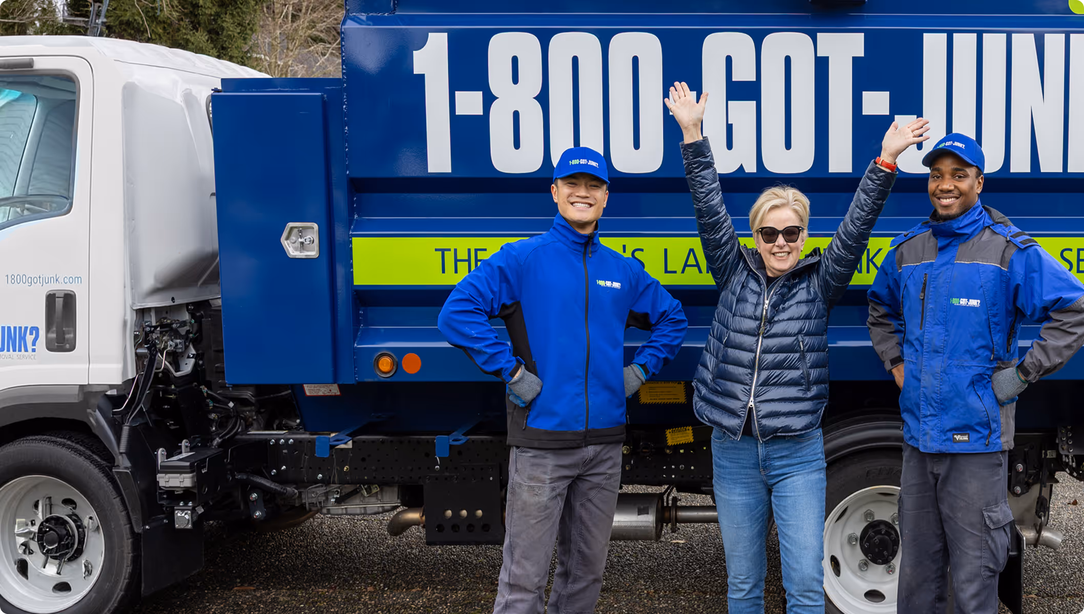 Three people smiling and giving thumbs up in front of a blue 1-800-GOT-JUNK? truck, two wearing blue uniforms and one in casual clothes.