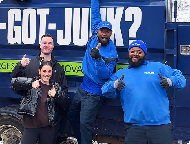 Four people smiling and giving thumbs up in front of a blue 1-800-GOT-JUNK? truck, two wearing blue uniforms and two in casual clothes.