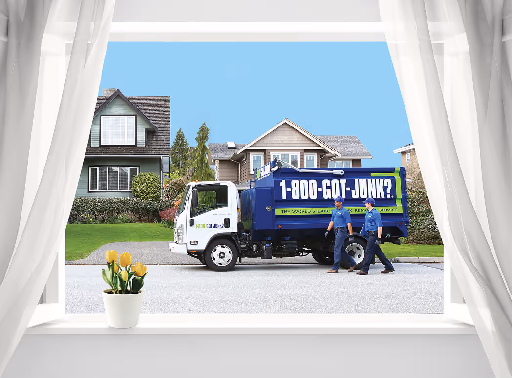 View through a window with sheer white curtains of a 1-800-GOT-JUNK? truck parked on a residential street with two workers walking beside it.