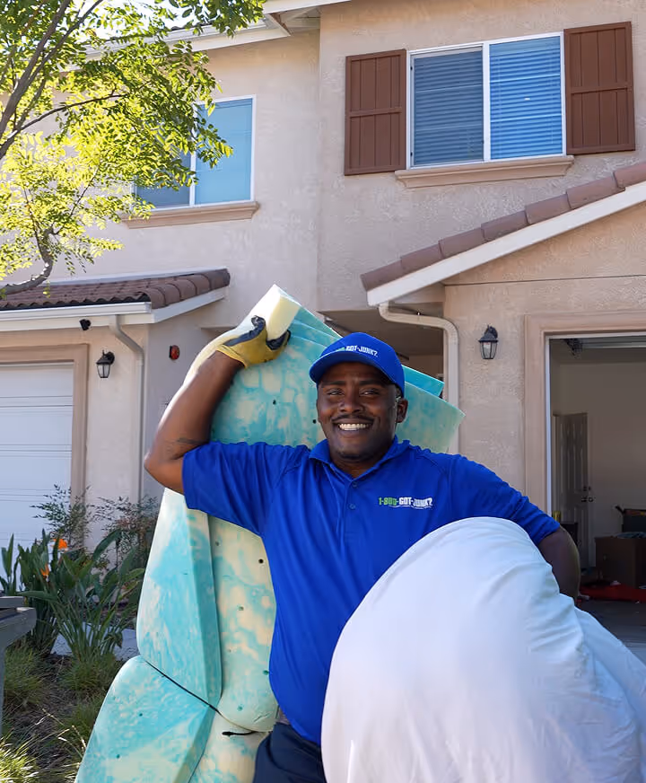 Smiling 1-800-GOT-JUNK? worker carrying a large foam mattress and pillow outside a suburban home during daylight.
