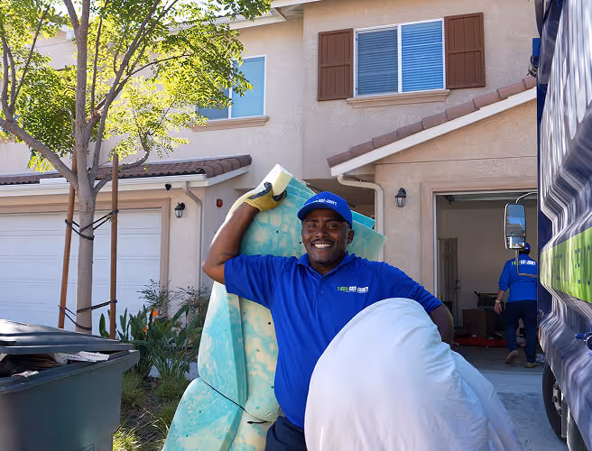 Smiling 1-800-GOT-JUNK? worker carrying a large foam mattress and pillow outside a suburban home during daylight.