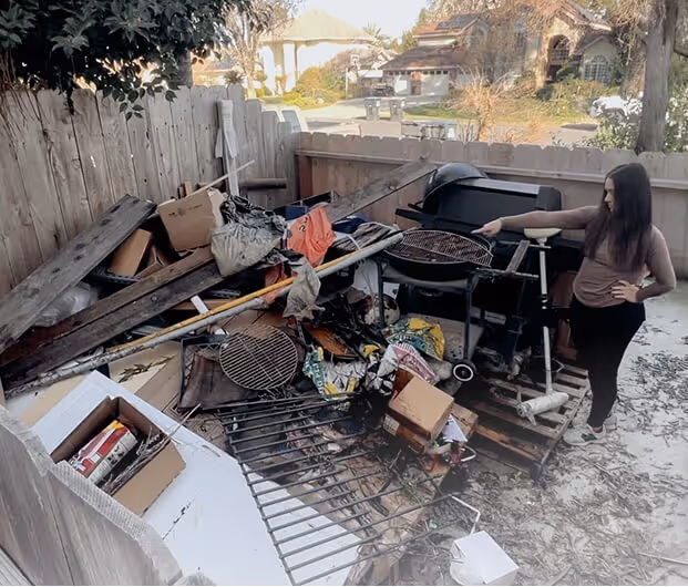 Woman standing next to a cluttered outdoor area filled with wooden pallets, metal grill parts, cardboard boxes, and various debris against a wooden fence.