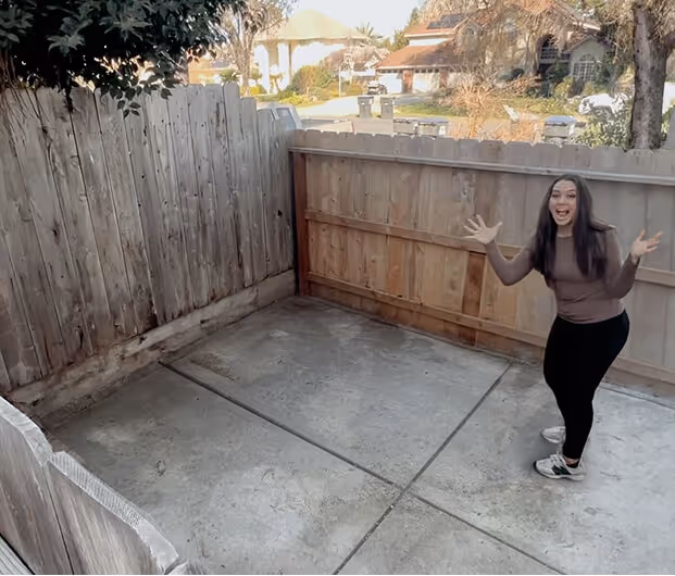 Woman smiling and gesturing excitedly in a small fenced concrete backyard.