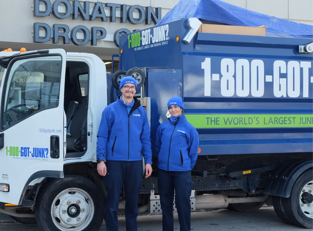 Two 1-800-GOT-JUNK? workers in blue jackets and hats stand beside their junk removal truck outside of a Donation Drop. 