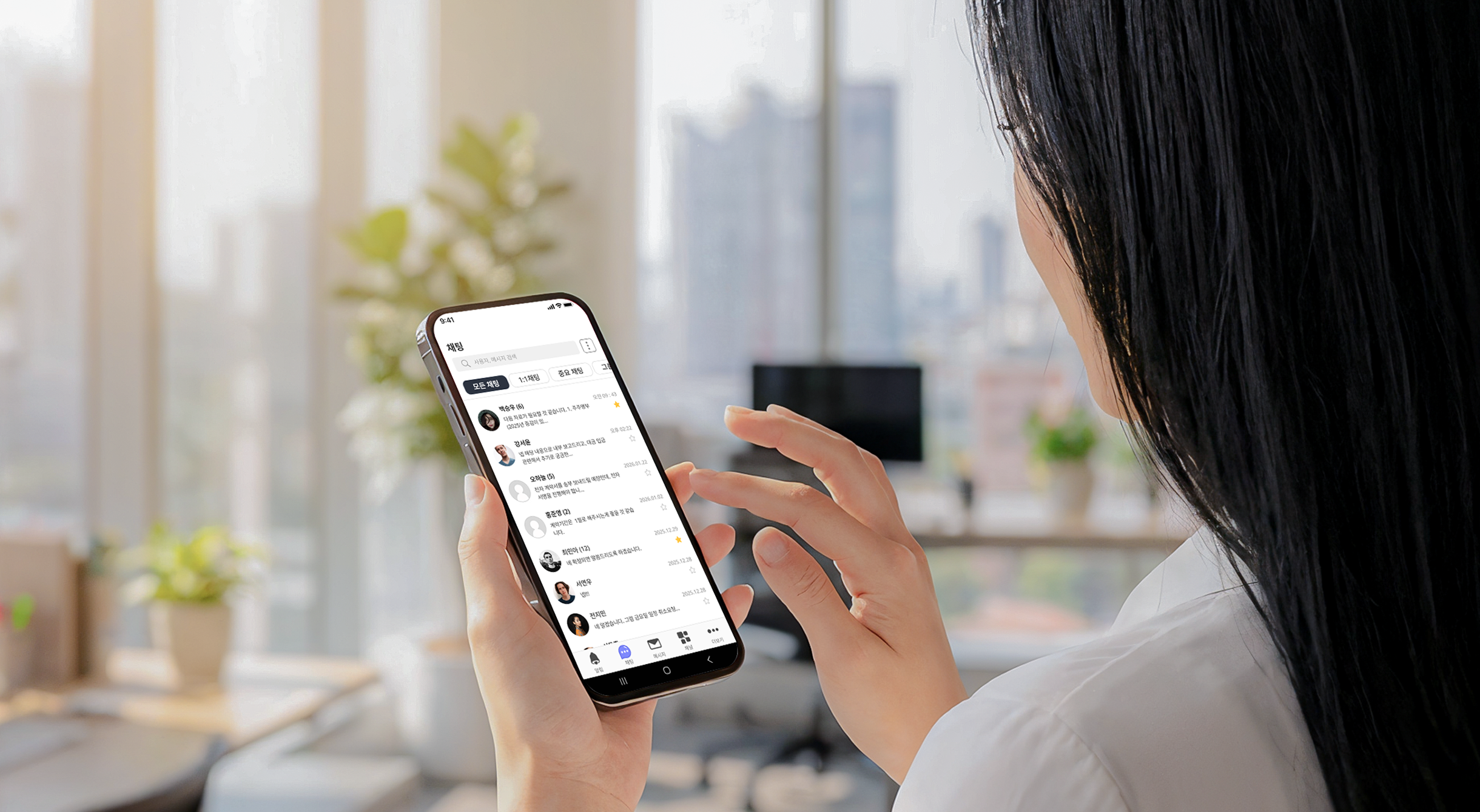 Person holding smartphone showing a Korean chat messaging app screen in a bright office setting.
