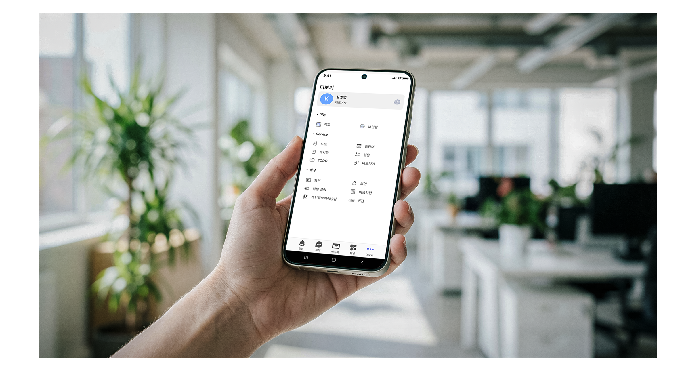 Hand holding a smartphone displaying a Korean menu interface with options for notes, calendar, security settings, and more in a bright office background.