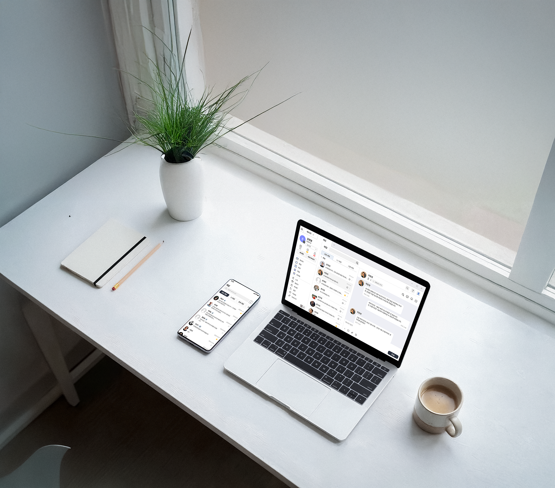 White desk with a laptop and smartphone displaying chat apps, a potted plant, notebook with pencil, and a cup of coffee near a window.