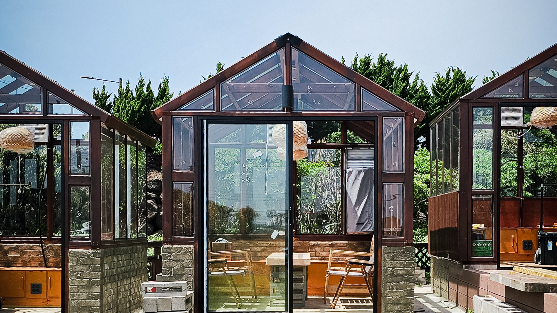 Three small glass greenhouses with brown frames, each containing wooden chairs and tables, under a clear blue sky.