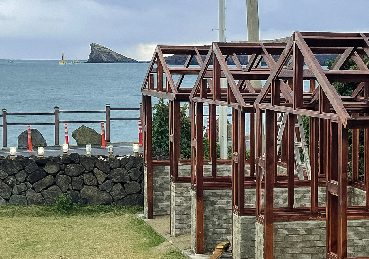 Wooden framework of small structures near a coastal stone wall with the ocean and a rocky island in the background.