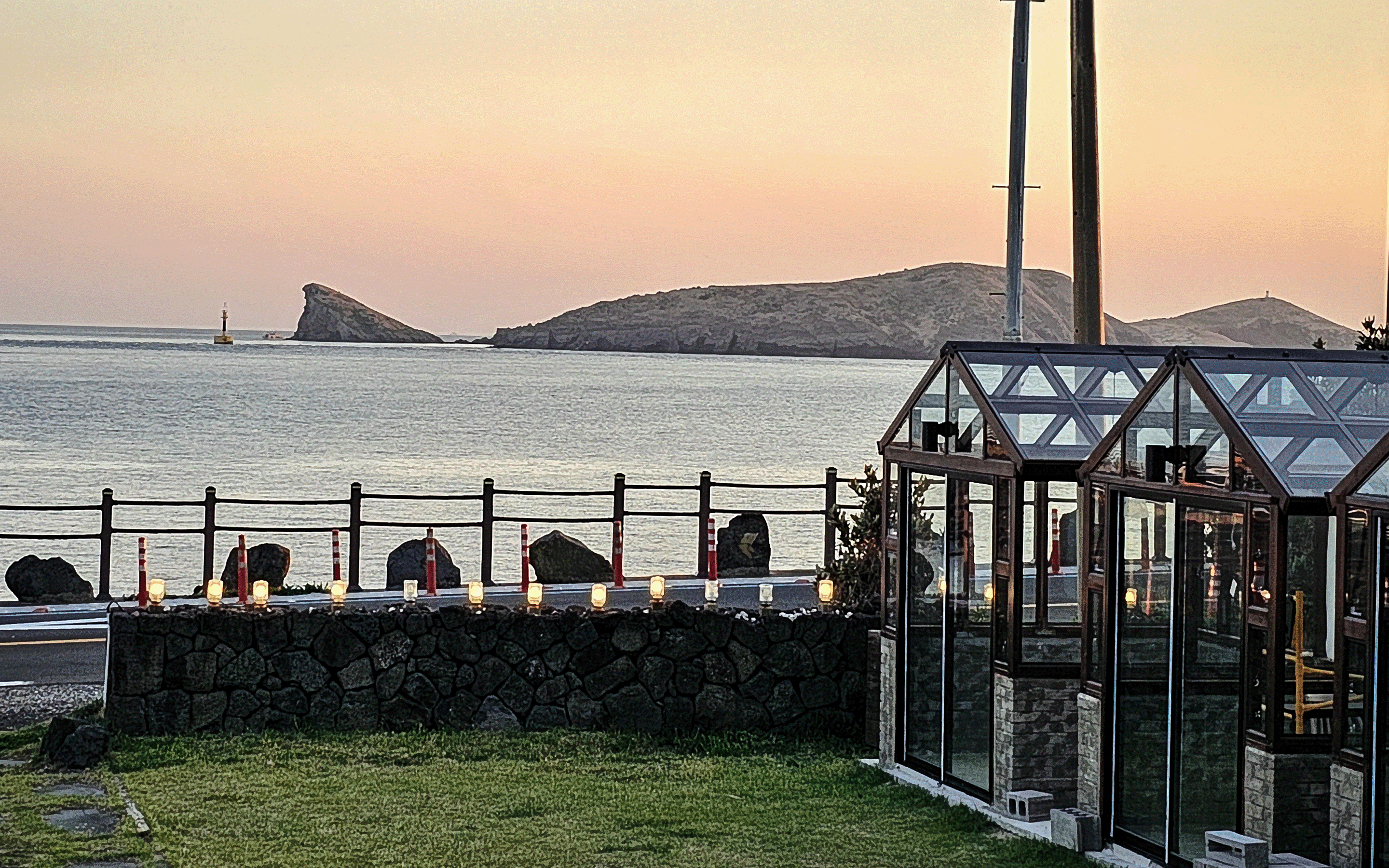 A glass greenhouse structure on a grassy area near a stone wall and road overlooking a calm sea with rocky islands at sunset.