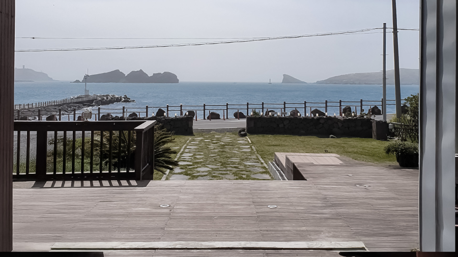 View through a glass door of a wooden deck and green lawn leading to a pier with a lighthouse, overlooking a calm sea with rocky islands in the distance.