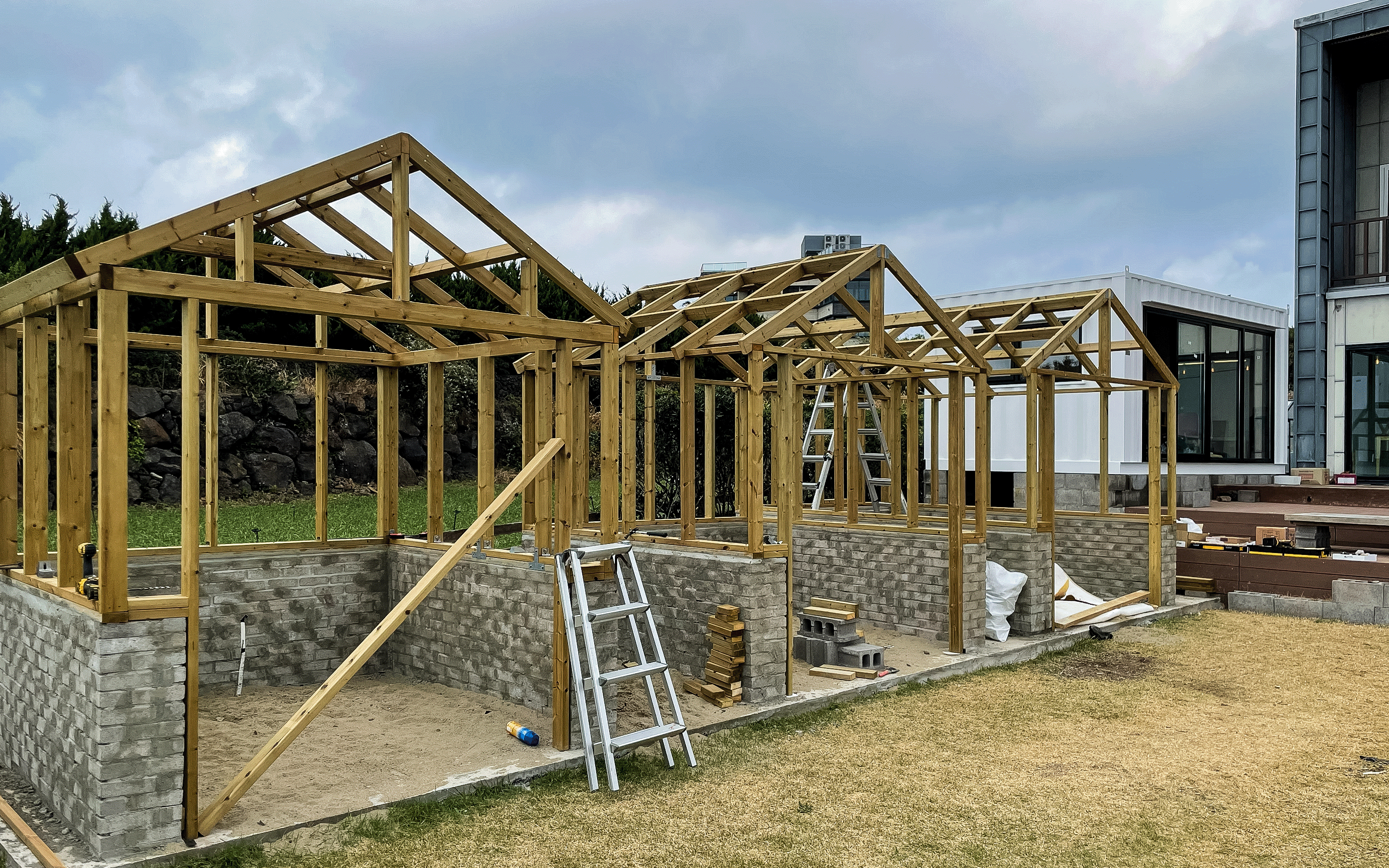 Three small greenhouses under construction with wooden frames and brick bases on a grassy plot beside a modern building.