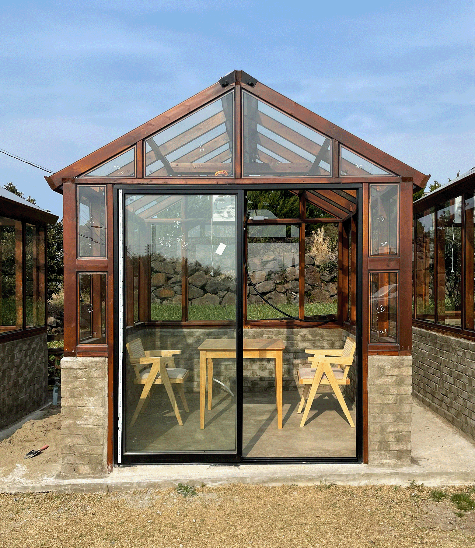 Small wooden greenhouse with glass walls containing a wooden table and two matching wooden chairs inside.