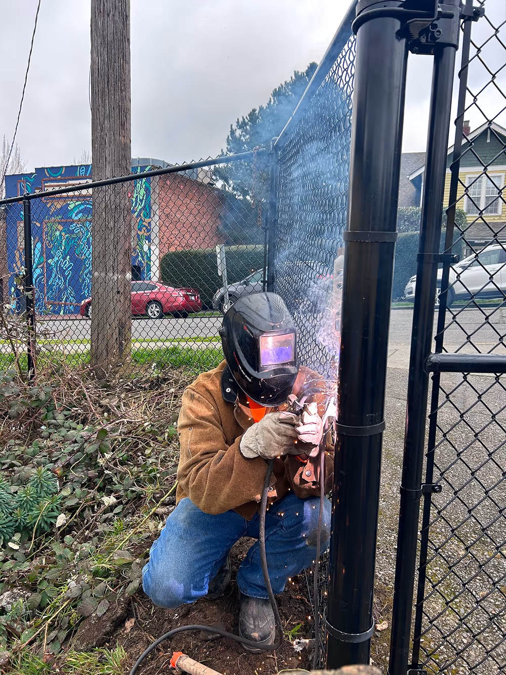 Person wearing a welding helmet and gloves kneeling and welding a black metal fence post outdoors.