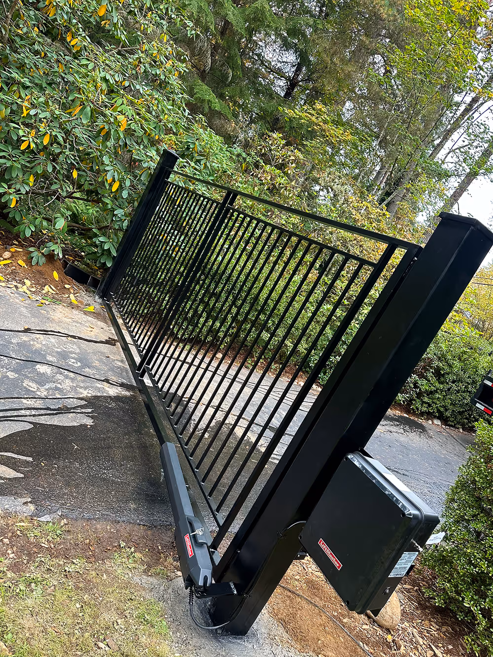 Black metal automatic driveway gate installed on a concrete driveway surrounded by green shrubs and trees.