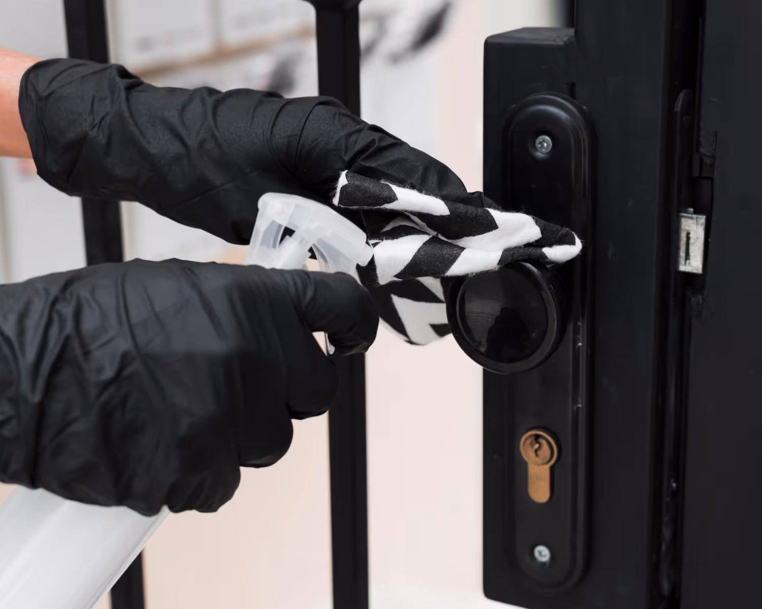 Person wearing black gloves cleaning a black door knob with a striped white and black cloth and spray bottle.