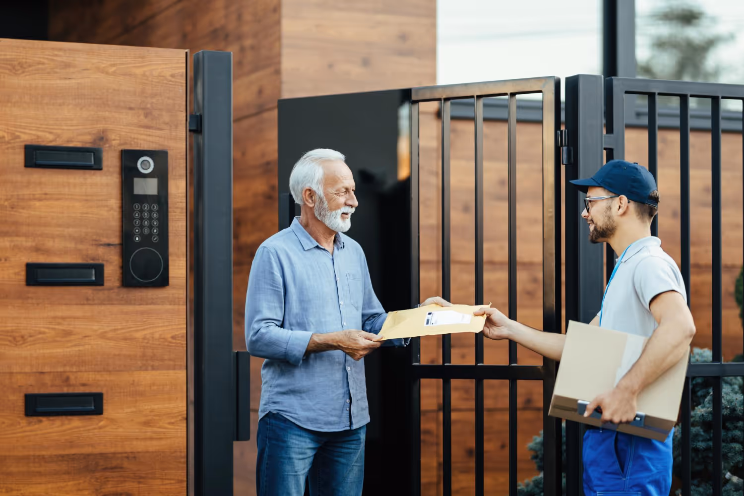 Delivery person handing a package to an elderly man at the gate of a modern wooden house.