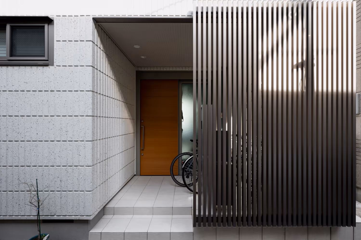 Modern home entrance with wooden door, white tiled steps, and a vertical metal slat privacy screen partially obscuring bicycles behind it.