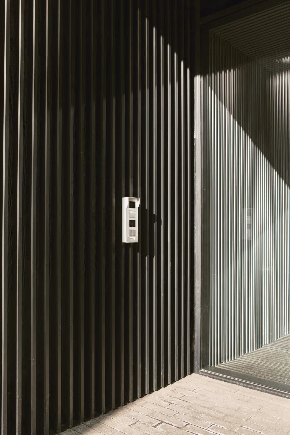 Modern black vertical slat wall with a white intercom system mounted near a glass corner with vertical blinds.