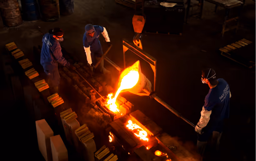 Três trabalhadores em uniforme derramando metal fundido incandescente em moldes numa fundição industrial.
