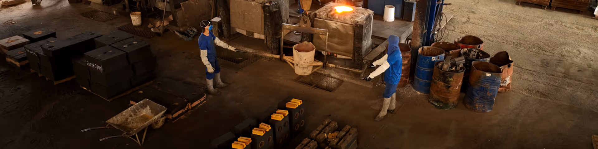Trabalhadores em uniforme azul e equipamentos de proteção manuseando fundição em uma fábrica industrial.