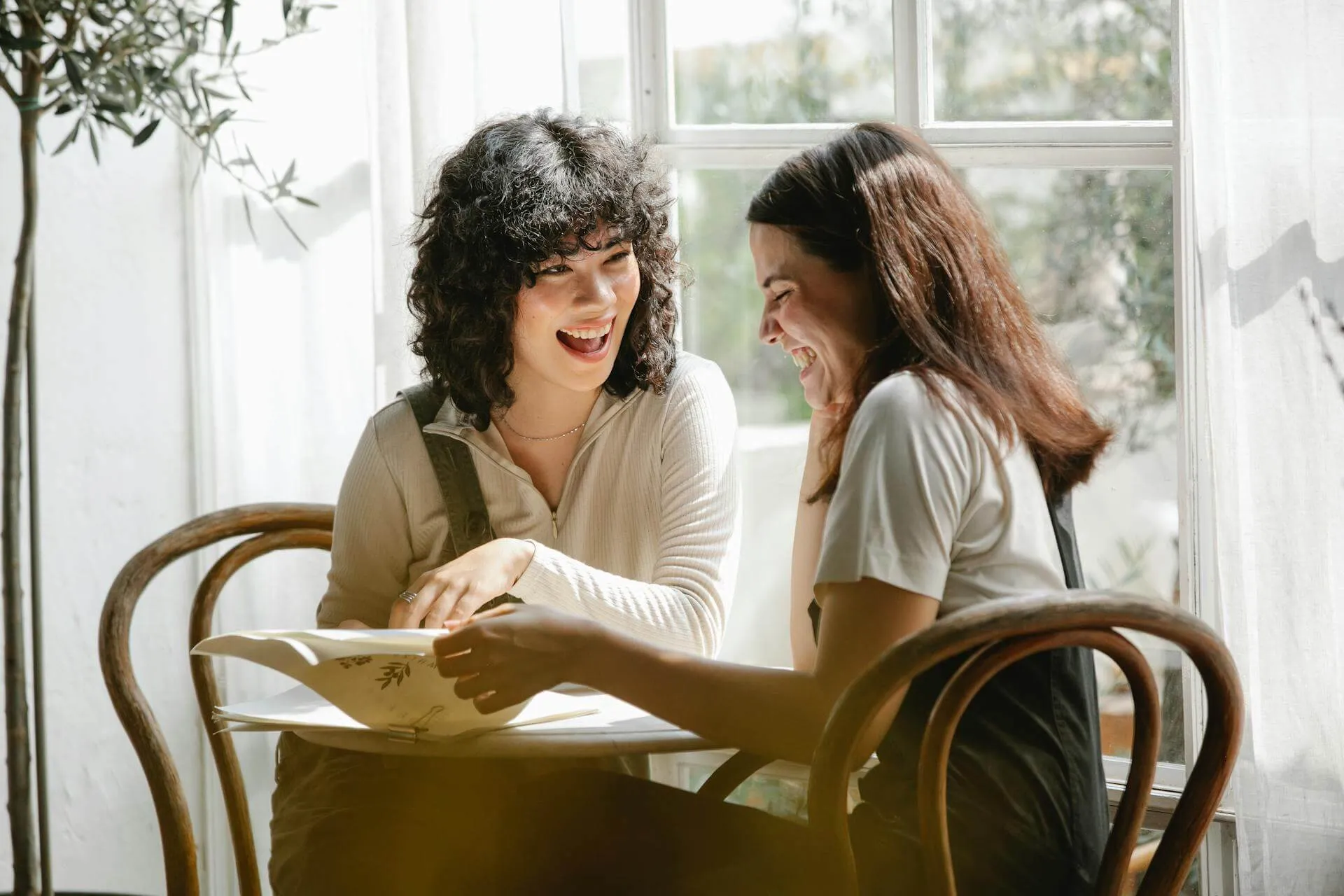 Two young women sitting at a table near a window, laughing and looking at a book together.