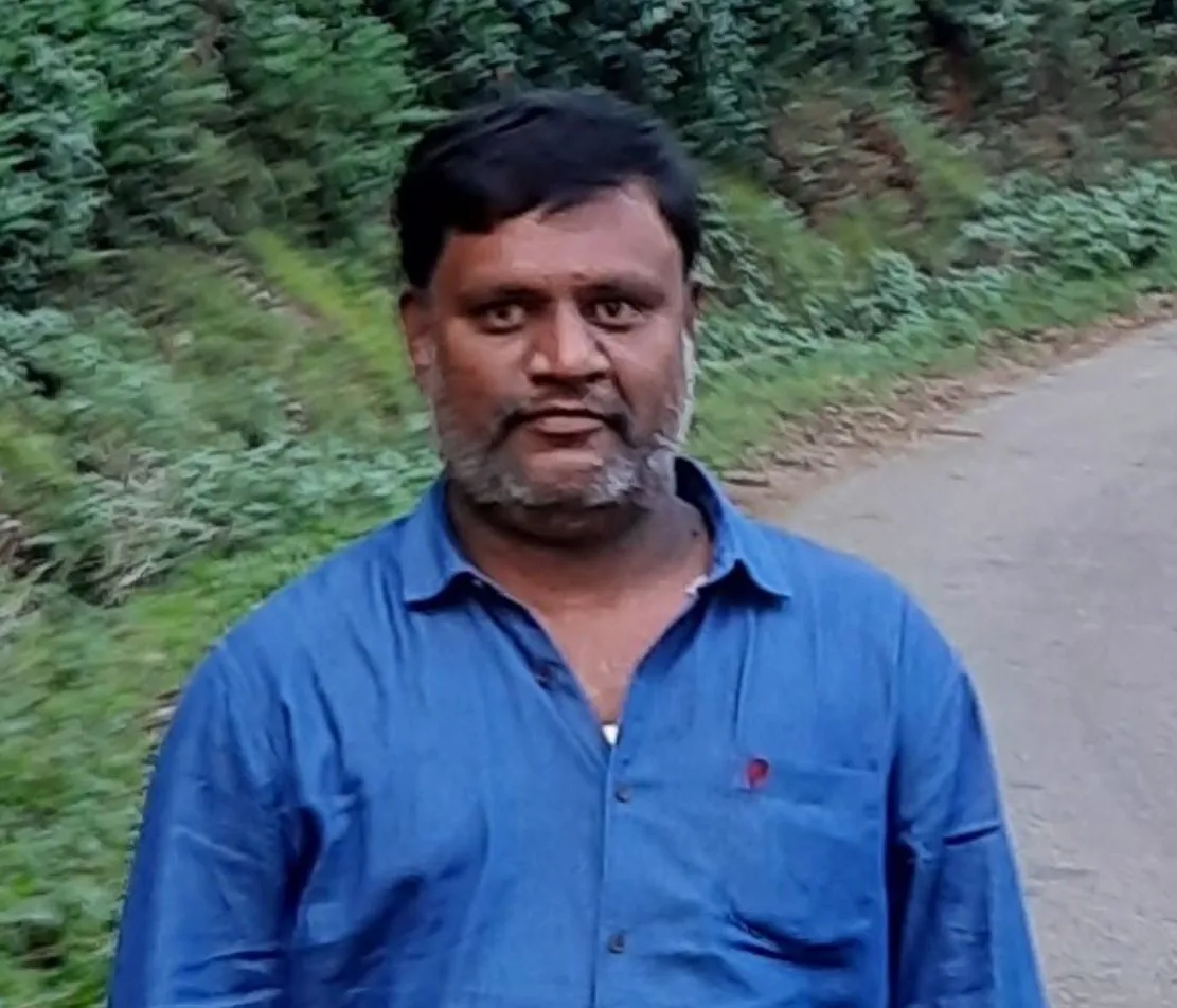 Man with short black hair and beard wearing a blue shirt standing outdoors near a green bushy area by a dirt path.