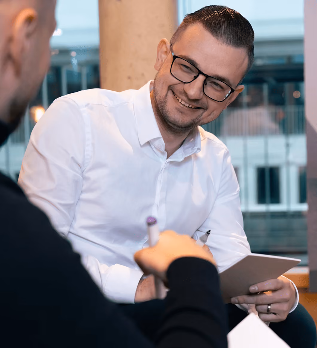 Ein Mann mit Brille und weißem Hemd sitzt in einer modernen Umgebung und lächelt im Gespräch. Er hält ein Tablet in der Hand, während ihm eine zweite Person mit Stift gegenübersitzt. Die Szene wirkt entspannt und kollegial.