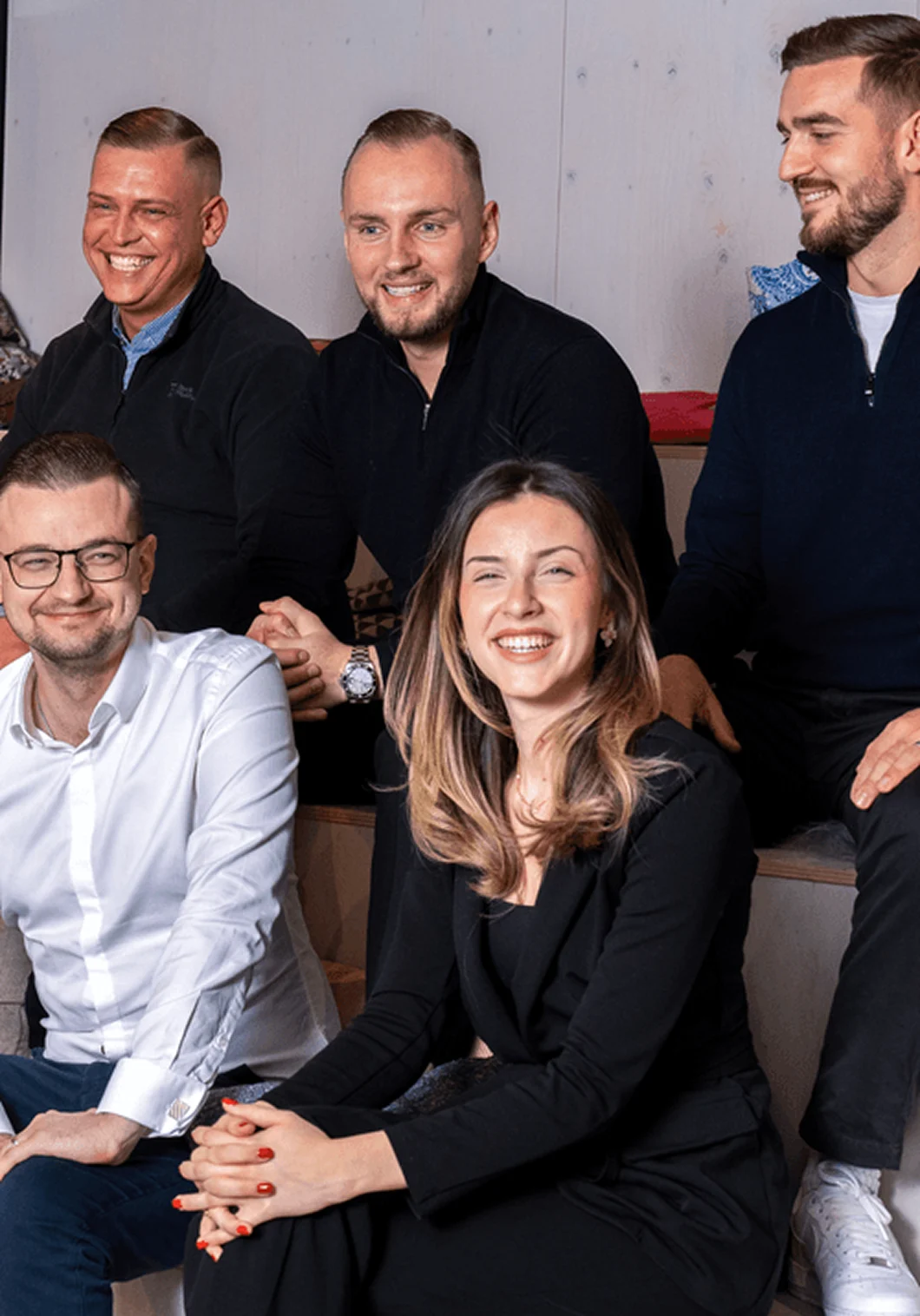 Sechs Personen sitzen locker auf Stufen und lachen gemeinsam in die Kamera. Die Szene wirkt freundlich und ungezwungen, mit moderner Büro-Atmosphäre im Hintergrund. Die Gruppe wirkt motiviert und gut gelaunt, als würde sie Teil eines eingespielten Teams sein.