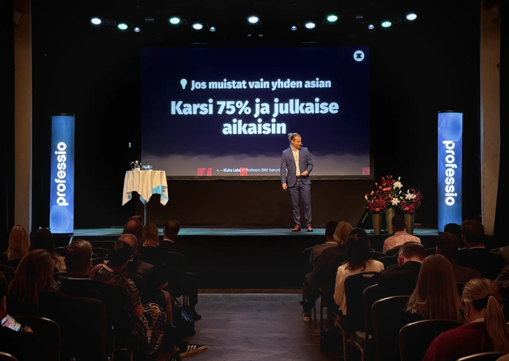 Antti Salonen puhuu lavalla yleisölle konferenssissa, taustalla suuri näyttö, jossa lukee: Jos muistat vain yhden asian, karsi 75% ja julkaise aikaisin.