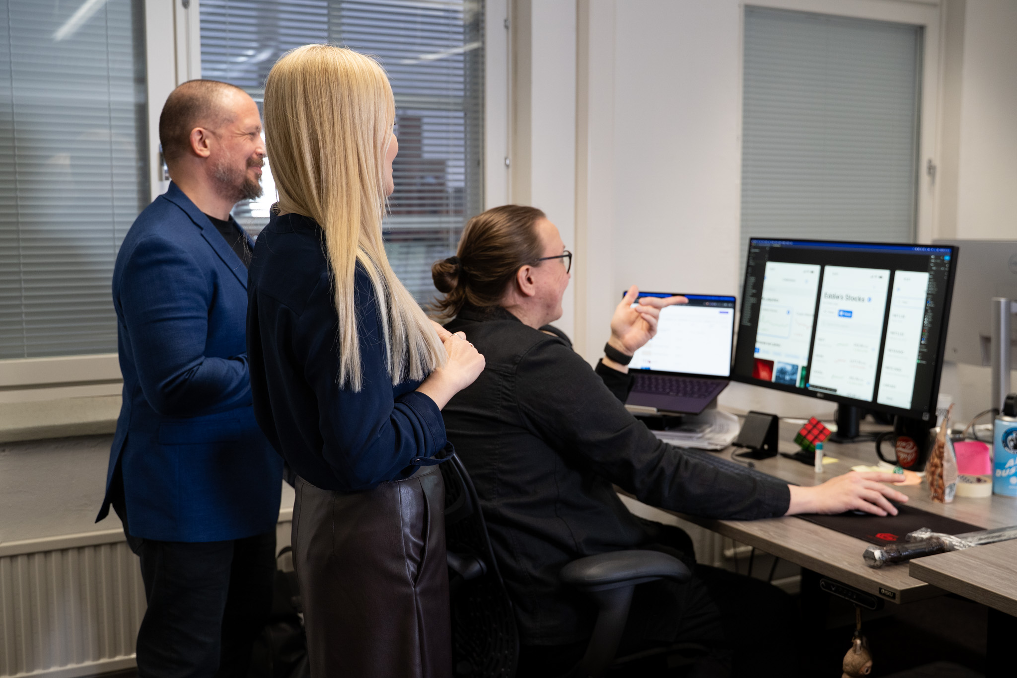 Kolme henkilöä työskentelee toimistossa, yksi istuu näyttöjen ääressä ja osoittaa tietokoneen näyttöä, kaksi seisoo hänen takanaan katsomassa näyttöä.